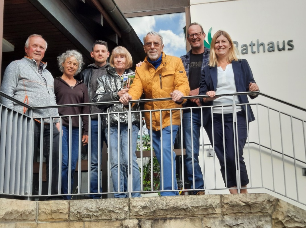 Auf dem Bild sind sieben Personen zu sehen. Links steht ein Herr mittlerer Größe in den 70er Jahren. Er hat graues kurzes Haar und einen grauen Fleecepullover an. Rechts daneben steht eine Frau, die ein bisschen kleiner ist als der neben ihr stehende Mann. Sie ist ca. 1,60 groß und hat eine braune Bluse und eine blaue JEans an. Sie hat graue krause Haare. Die dritte Person von Links ist ca. 30 Jahre alt. Der Mann hat einen Kapuzenpullover und eine Lederjacke an. Er hat kurze dunkle Haare. In der Mitte steht eine Frau mit kinnlangem Haar. Sie ist leger gekleidet. Rechts daneben steht ein älterer Herr im alter um die 80 Jahre. Er hat eine gelbe Jacke und eine hellblaue Jeans an. Er stützt sich auf dem Geländer vor sich mit beiden Händen ab, den einen Fuß deutlich vor den anderen gestellt. Er hat einen weißen kurzgehaltenen Bart und kurzes graues Haar. Zudem trägt er eine Brille, die durch die Lichteinstrahlung leicht bräunlich verfärbt ist. Rechts neben dem Herrn steht ein weiterer Herr. Er ist die zweite Person von rechts etwas hinter seinen nachbarn gestellt. Er blickt über die Schultern der beiden. Er hat eine Jeansjacke an und ist über 1,85m groß. Er hat kurzes Haar und eine Brille auf. Die rechte Person ist eine Frau mit schulterlangem blonden Haar. Sie stützt sich ebenfalls auf dem Geländer auf. Sie hat einen blauen Blazer und eine dunkelblaue 7/8 Hose an. Darunter trägt sie eine weiße Bluse. Die Dame lacht sehr freundlich. Sie ist die Bürgermeisterin Catenazzo. Alle stehen auf einer Anhähe. Rechts scheint eine Treppe zu beginnen. Im Hintergrund ist ein Gebäude zu sehen. Es steht ein Schriftzug mit "Rathaus" darauf.