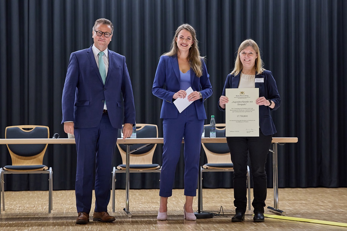 Es stehen drei Personen in Anzug vor einem schwarzen Vorhang. Direkt hinter den drei Personen stehen Tische mit Stühlen. Der Boden ist in einem hellen Holzton gehalten. Die linke Person hat einen blauen Anzug und eine türkise Krawatte an. Der Mann hat kurzes leicht graues Haar und ist um die 60 Jahre alt. In der Mitte und rechts stehen zwei Frauen mit langen Haaren. Die mittlere Dame hat ebenfalls einen blauen Anzug an, der sportlich geschnitten ist. Dazu trägt sie rosa Schuhe und ein hellblaues Oberteil. Sie hat lange braune Haare, die auf der Brust enden. Die Haare sind offen getragen. In der Hand hält sie weiße Moderationskarten. Die Rechte Frau (Bürgermeisterin Catenazzo) trägt einen dunkelblauen Blazer, eine schwarze Hose und eine beige Bluse. Ihr Haar ist blond, schulterumspielend und offen getragen. In beiden Händen hält sie eine große Urkunde.