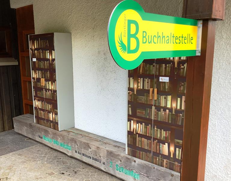 Ein großes Schild in grün-gelb mit der Aufschrift "Börtlinger Bücherhaltestelle" hängt vor zwei großen Schränken, die mit Büchern bedruckt sind.