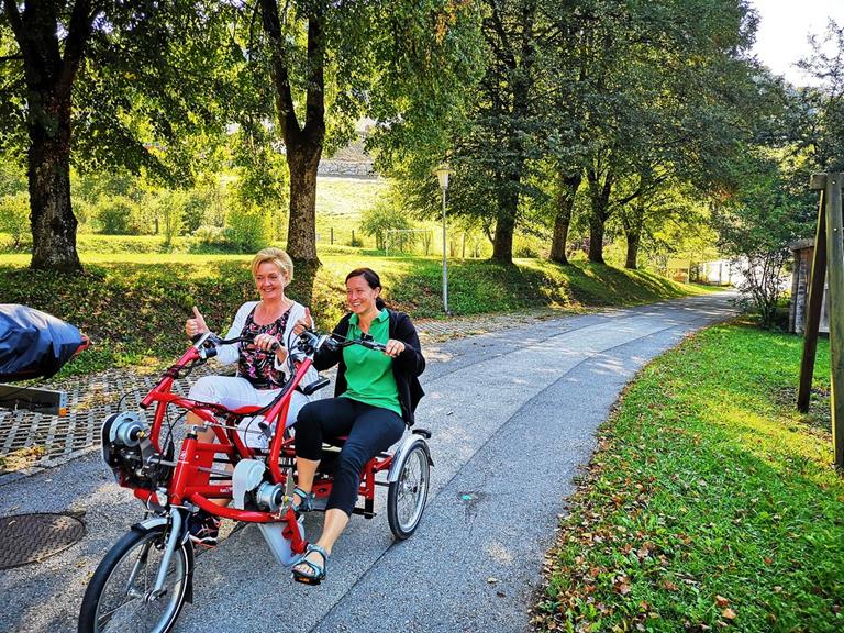 Zwei Damen mittleren Alters sitzen auf einem roten Paralleltandem und fahren auf einem schmalen asphaltierten Weg unter Bäumen. Beide sehen glücklich aus und lächeln.